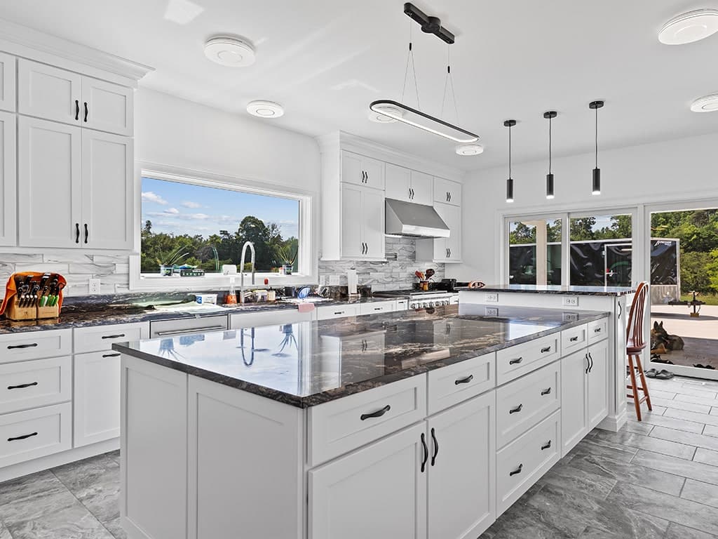 Modern kitchen with white cabinets, a large island with dark countertops, stainless steel appliances, pendant lights, and large windows overlooking a patio and greenery outside.