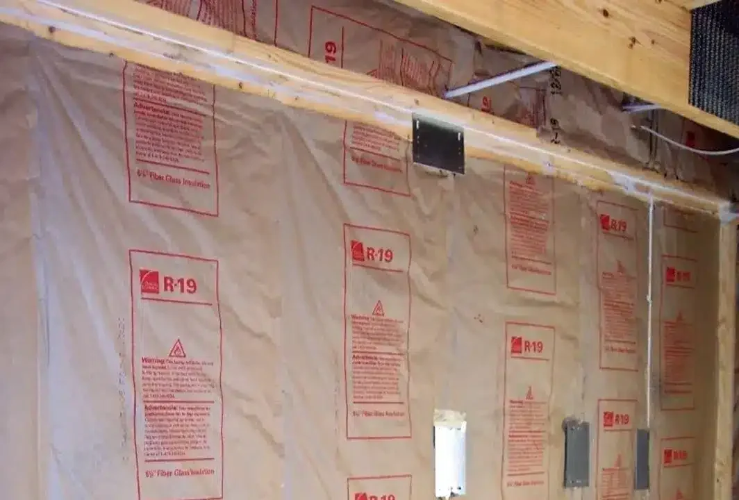 A wall with R-19 fiberglass insulation installed between wooden studs, covered with kraft paper facing. Electrical boxes and wiring are visible, indicating ongoing construction or renovation work.