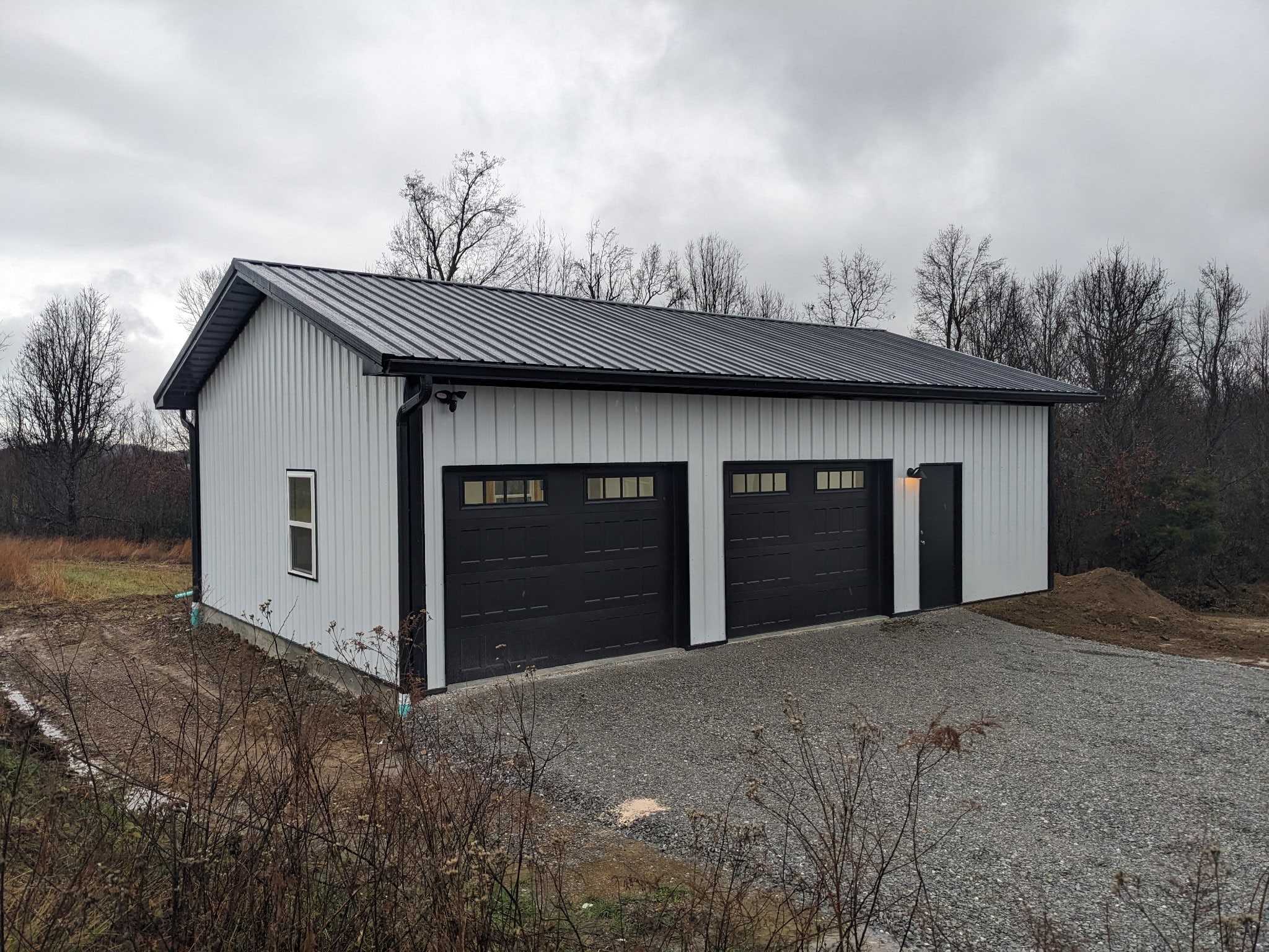 A white metal garage building with a black roof and three black garage doors, set on a gravel driveway. Surrounding area is grassy with some bare trees under a cloudy sky.