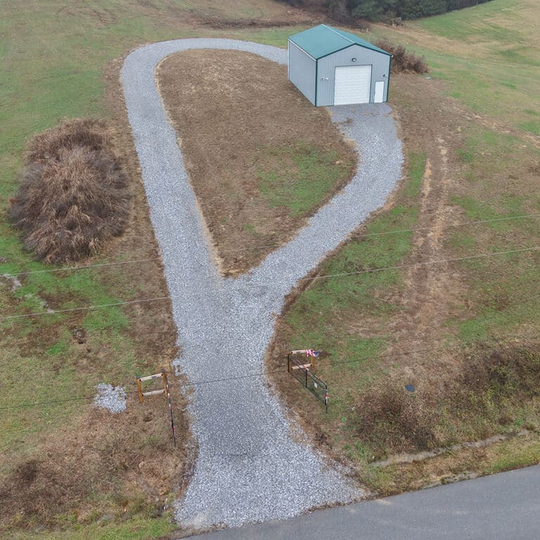 A gravel driveway shaped like a loop leads to a small green-roofed shed with a white door, surrounded by grass and a few bushes, next to a paved road—an inviting example of how Post-Frame Buildings can seamlessly blend into their surroundings.