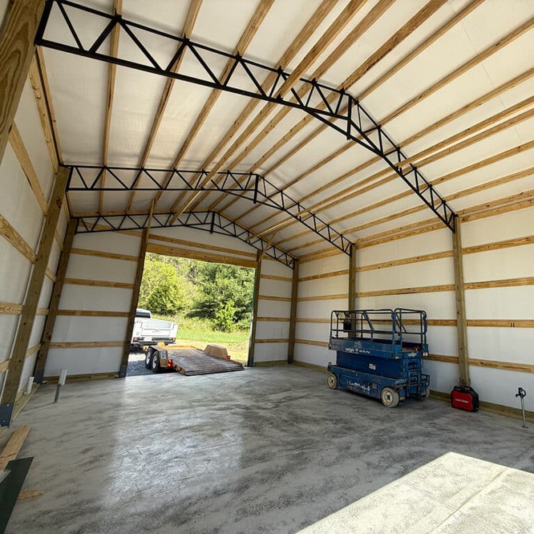 Interior of a large, unfinished metal building with exposed beams, highlighting the sturdy construction typical of Post-Frame Buildings. A blue scissor lift is inside, and a large open doorway reveals a truck and trees outside.
