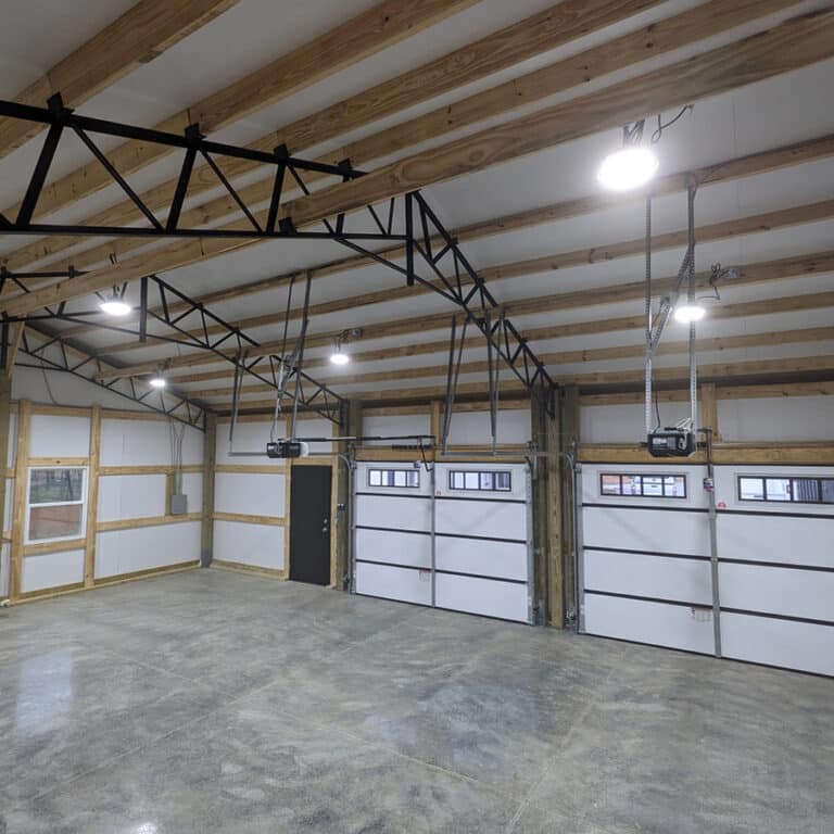 Spacious garage with three white overhead doors, exposed wooden beams, black metal trusses, bright ceiling lights, and polished concrete floor. Designed in the style of modern post-frame buildings, a black door and windows line the back wall.