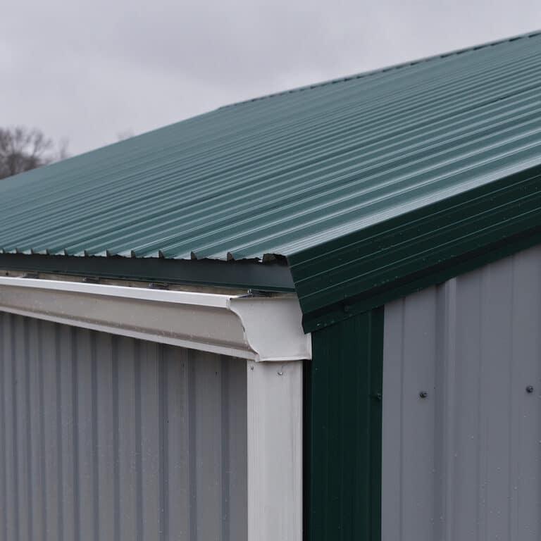Close-up of a green metal roof and gray metal siding on a post-frame building, showing overlapping panels and a white rain gutter at the roof edge under a cloudy sky.