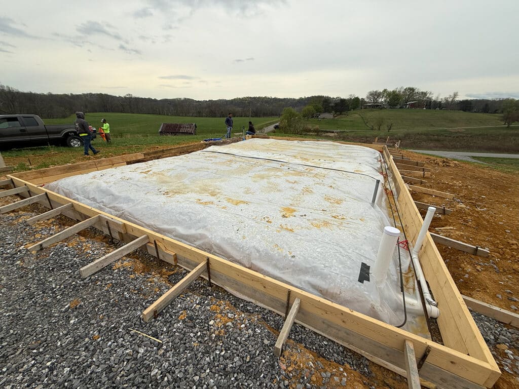 A construction site with a rectangular foundation covered in plastic sheeting, surrounded by wooden framing typical of Post-Frame Buildings, along with gravel and pipes, with workers and vehicles visible in the background.