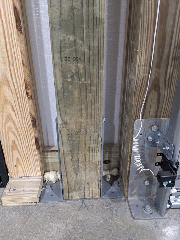 Close-up of three wooden beams anchored to a concrete floor with metal brackets and bolts, showcasing construction details typical of Post-Frame Buildings; expanding foam is visible at the base, and a white cord runs along one beam.