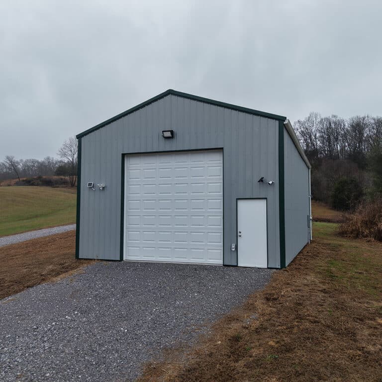 A gray metal post-frame building with a large white garage door and a small entry door, surrounded by grass and trees under a cloudy sky.