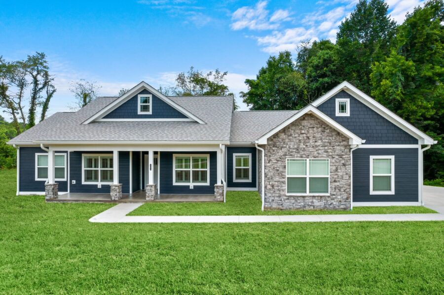 Single-story house with dark blue siding, white trim, stone accents, and a covered front porch. The home is surrounded by green grass and trees, with a driveway on the right. The sky above is partly cloudy.