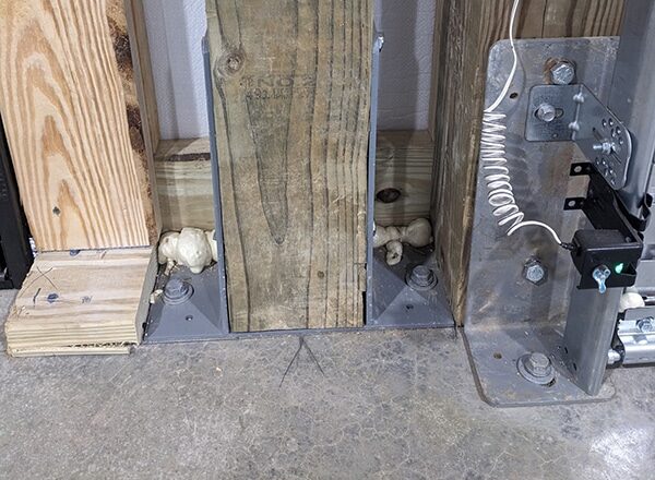 Close-up of wooden beams anchored with metal brackets to a concrete floor, with foam sealant filling gaps at the base and a coiled white cable nearby.