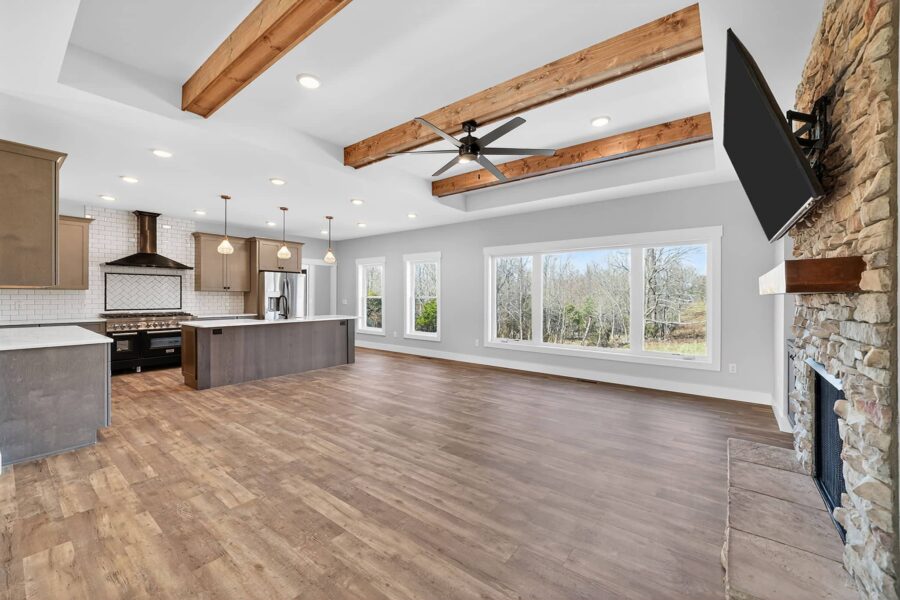 Spacious open-concept living area with wood floors, exposed ceiling beams, stone fireplace, large windows, modern kitchen with island, pendant lighting, and a wall-mounted TV.