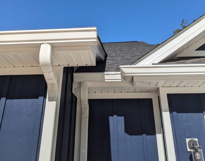 Close-up of beige rain gutters and downspouts attached to the blue exterior of a house, with a clear blue sky in the background.