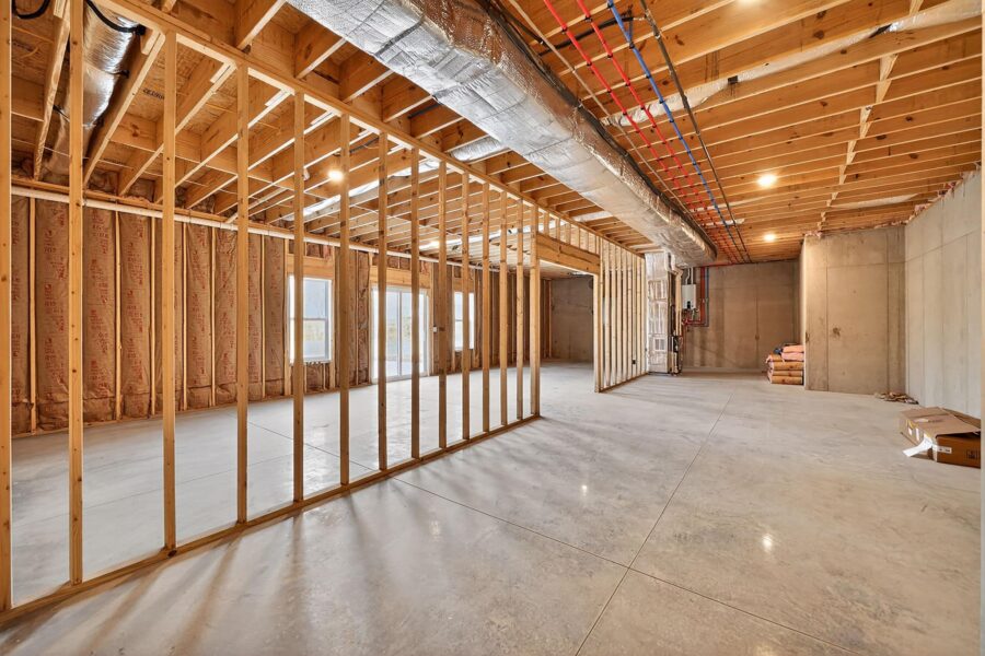 Unfinished basement with exposed wooden framing, insulation, ductwork, and concrete floors and walls. Large windows provide natural light. Construction materials are stacked in the background.
