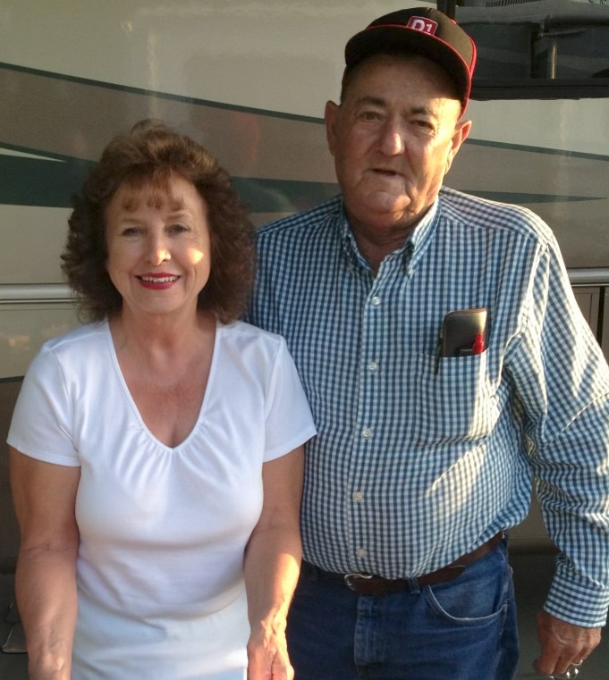 A woman in a white shirt stands smiling next to a man in a checkered shirt and baseball cap. They are outside, possibly near a vehicle or trailer.