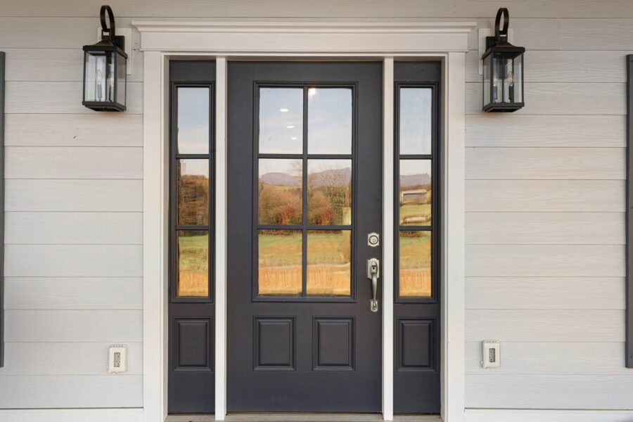 A black front door with rectangular glass panels is flanked by two narrow windows and lantern-style lights. The door reflects an outdoor landscape with trees and hills.