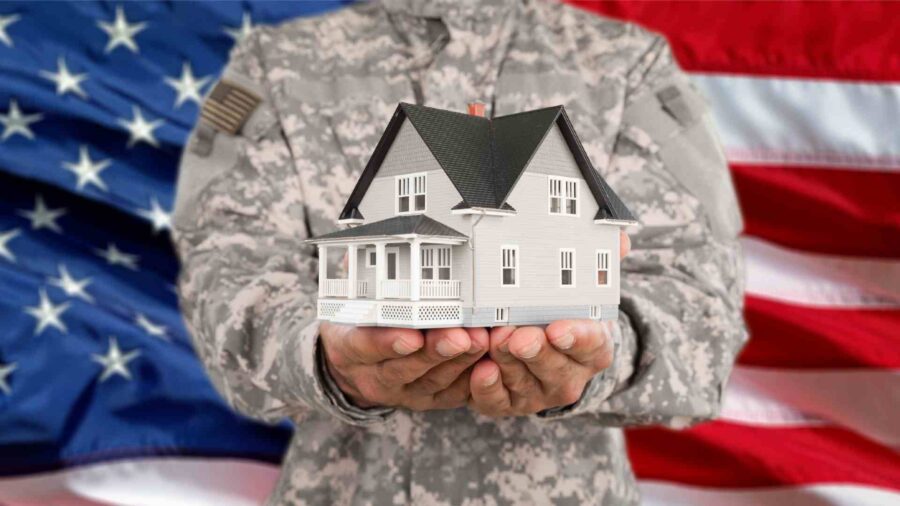 A person in military uniform holds a model house in their hands, with an American flag in the background.