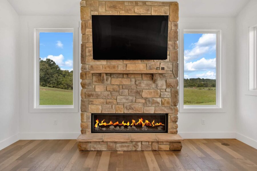 Stone fireplace with a mounted TV above, flames visible in a horizontal firebox, hardwood floor, and two windows showing green fields and blue sky with clouds.