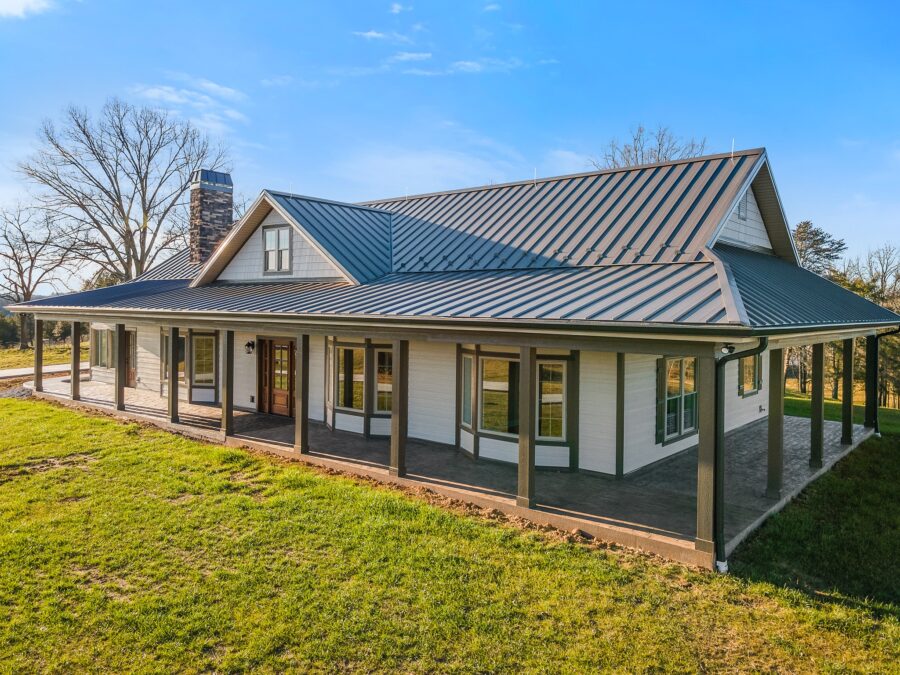 Single-story modern farmhouse with a metal roof, wraparound porch, large windows, and green lawn under a clear blue sky.