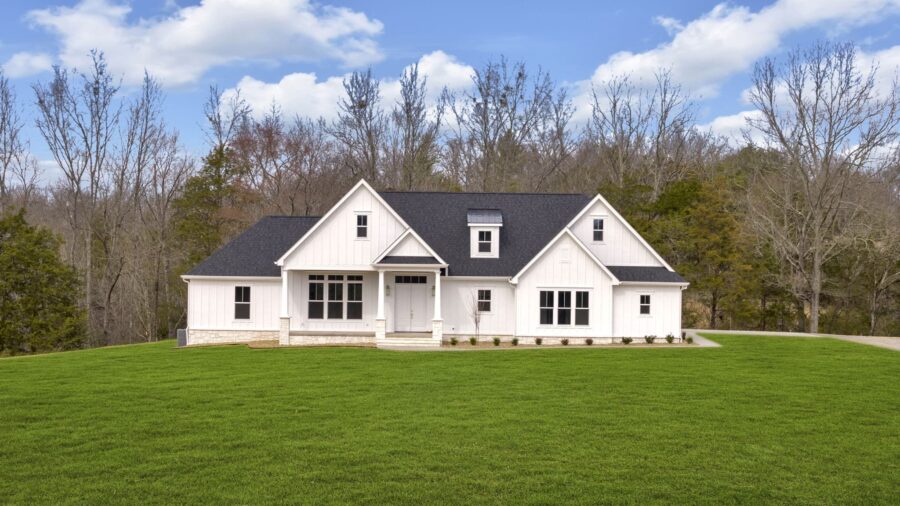 A white modern farmhouse with a dark roof sits on a large, neatly mowed green lawn. Bare trees and a blue sky with scattered clouds are in the background.