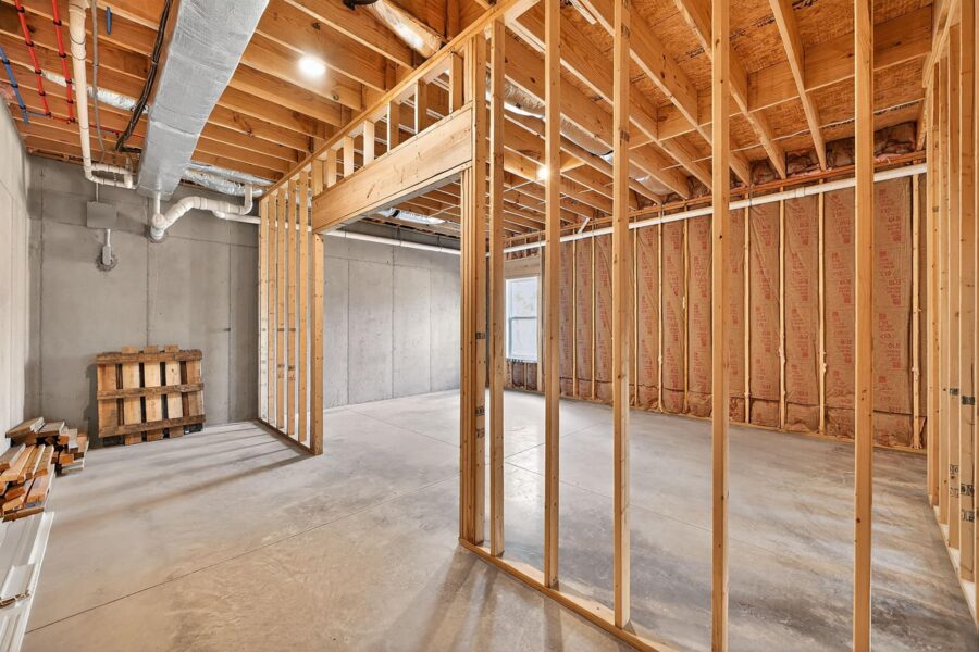Unfinished basement with exposed wooden framing, concrete floors and walls, visible insulation, and ductwork on the ceiling. A pallet and wood planks are stacked in one corner, and sunlight enters through a small window.