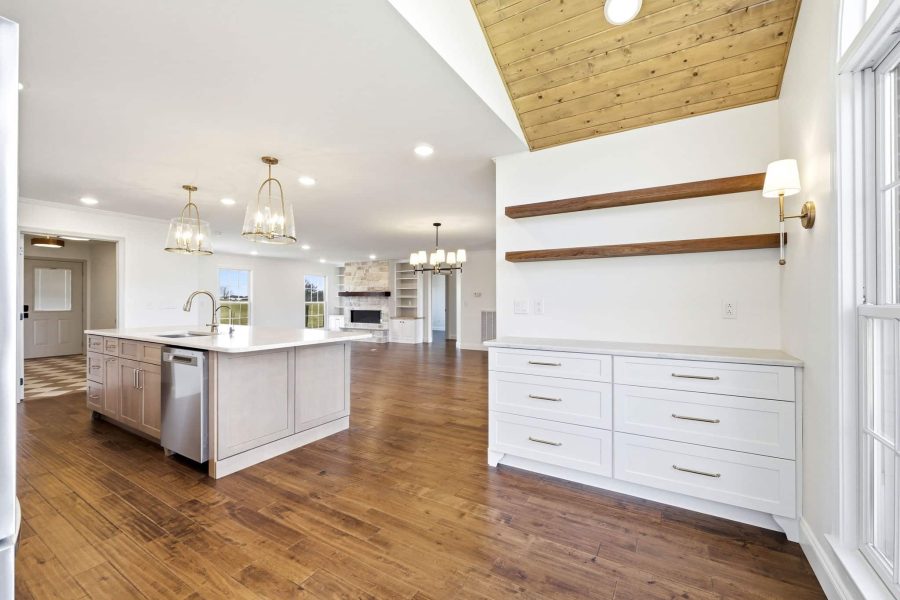Modern kitchen with wood floors, a large island with sink, white cabinets, open shelving, pendant lights, and a sloped wood ceiling accent. A living area with a fireplace is visible in the background.