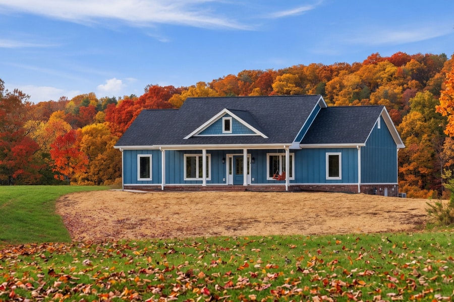 A blue house with white trim sits on a grassy lawn, surrounded by autumn trees with vibrant red, orange, and yellow leaves under a blue sky with clouds—a picturesque scene that invites you to slow down and relax, far from the rush of any busy schedule.