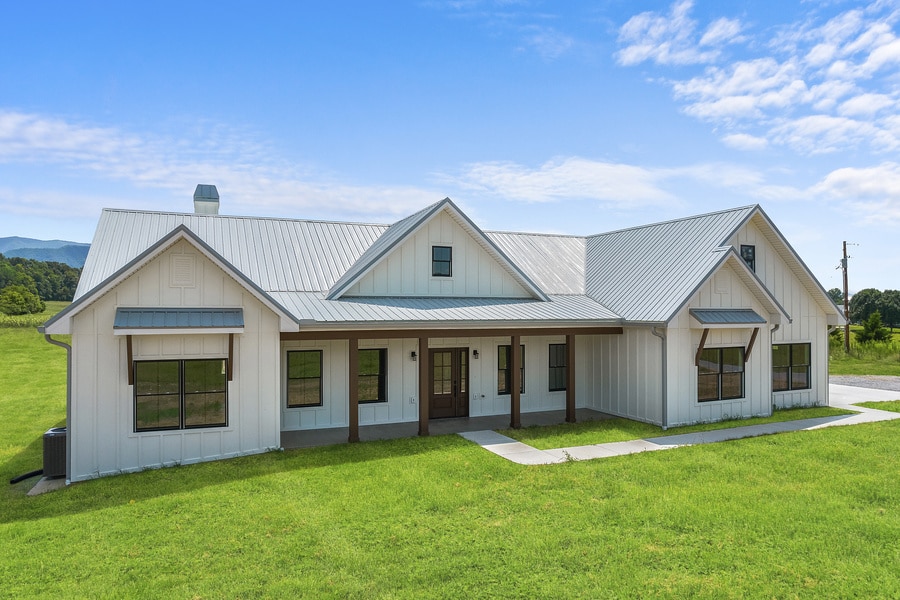 Modern white farmhouse with a metal roof, large windows, and a covered front porch—expertly crafted by a custom builder—set on a grassy lawn under a blue sky with scattered clouds.