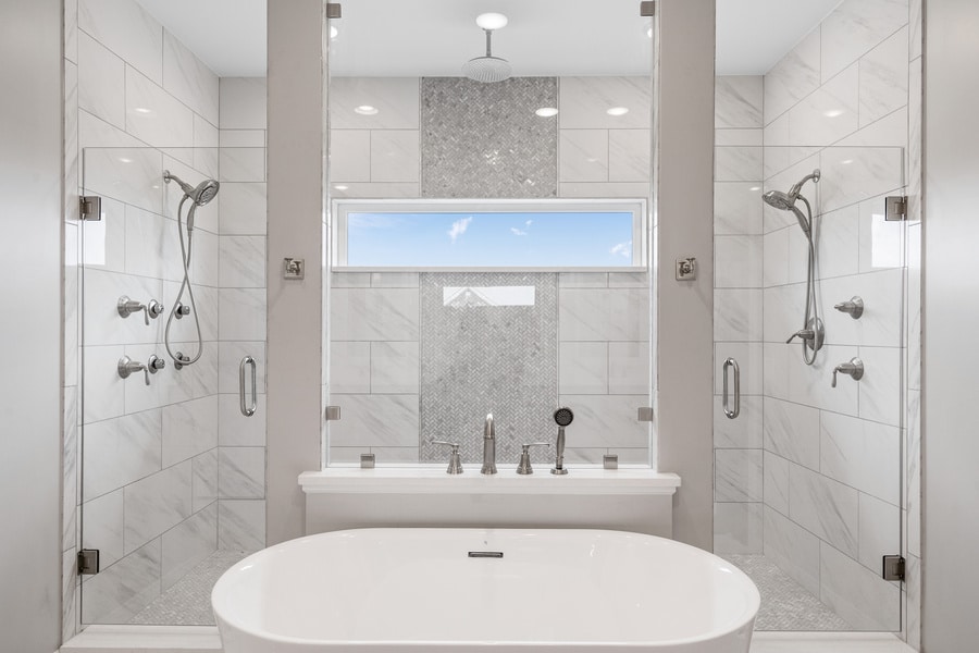 Modern bathroom with a white soaking tub, two glass-enclosed showers on either side, rainfall showerheads, white tile walls, and a window above the tub letting in natural light.